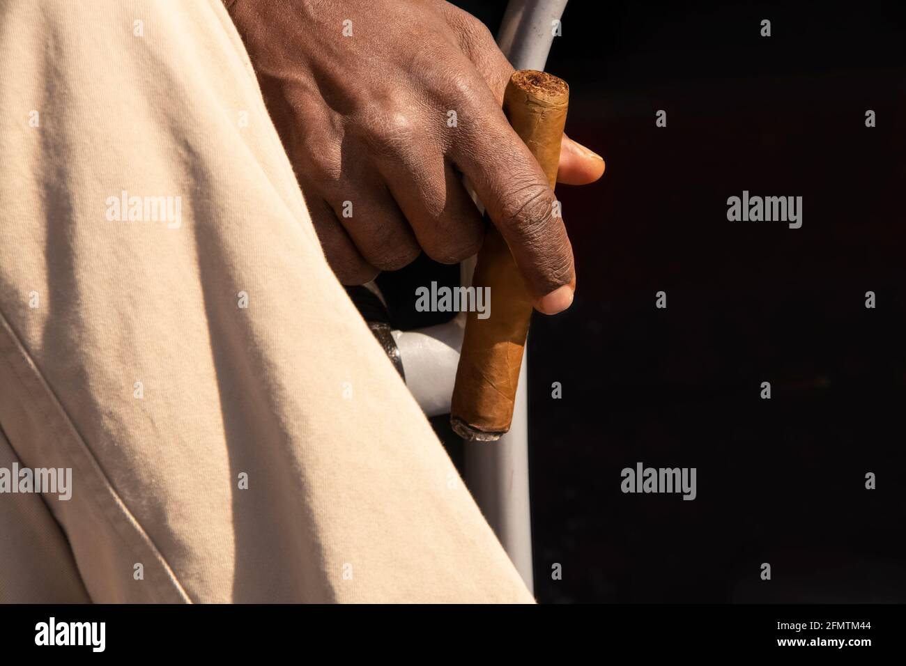 Das tropische Gefühl - Nahaufnahme einer beleuchteten Zigarre, die in der Nähe gehalten wird Zurücksitzender Mann unten durch gekreuztes Bein in hellem khaki hose Stockfoto