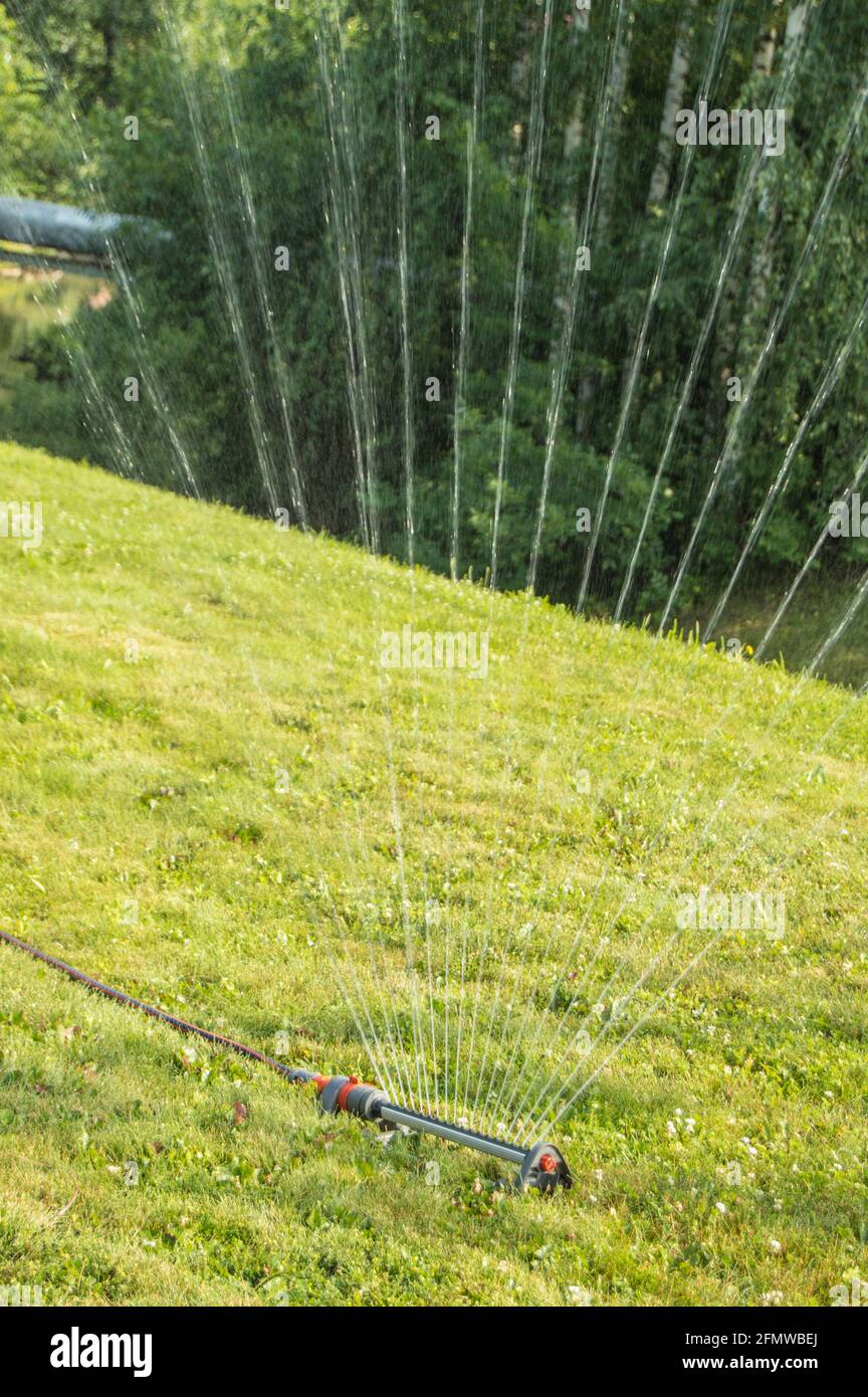 Ein Rasensprenger sprüht Wasser auf das grüne Gras, das am Hang wächst. Modernes Bewässerungssystem an einem sonnigen Sommertag im Freien. Stockfoto