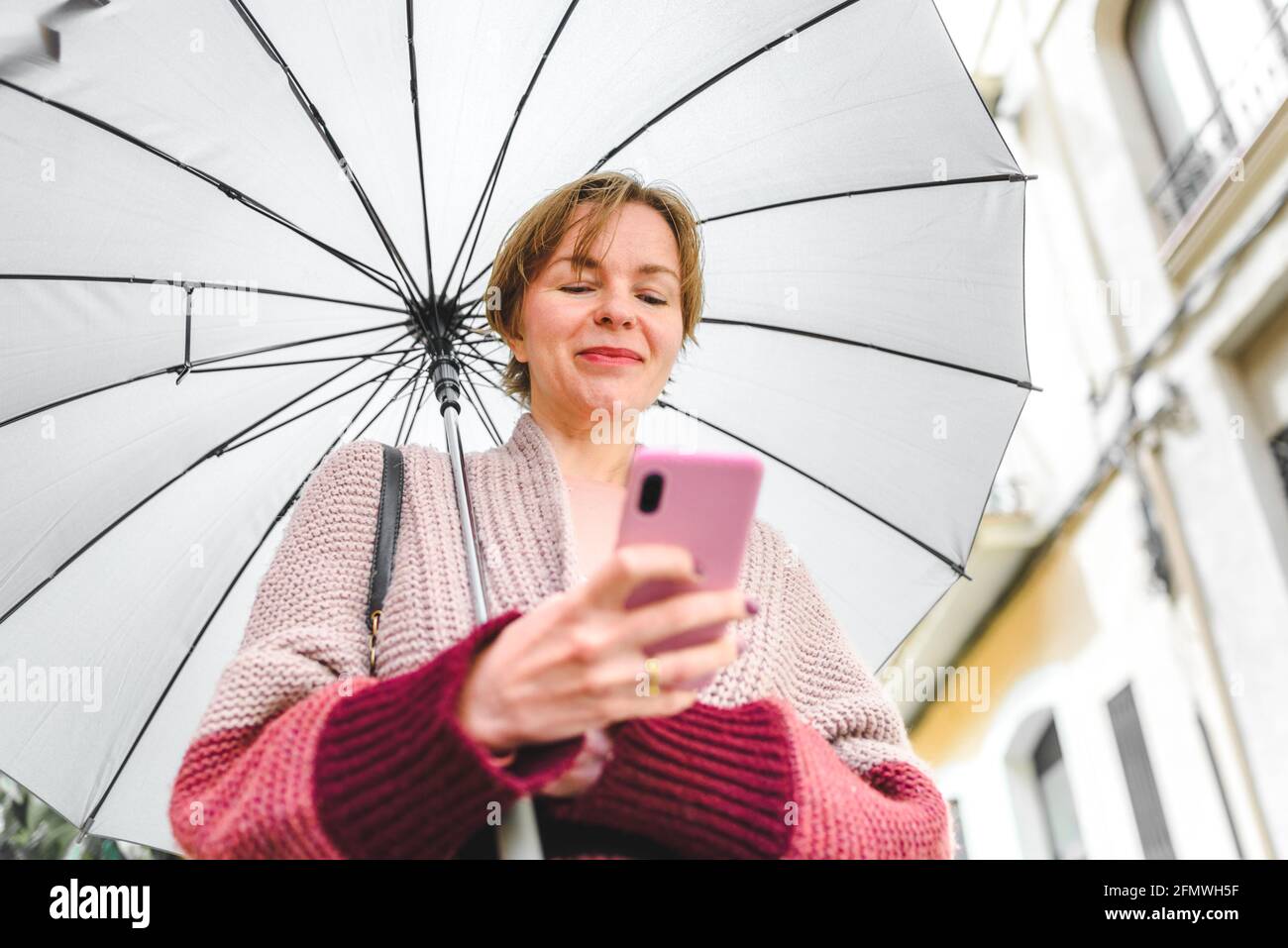 Horizontales Porträt einer blonden kaukasischen Frau, die ihr Telefon benutzt. Sie ist auf der Straße und hält einen Regenschirm Stockfoto