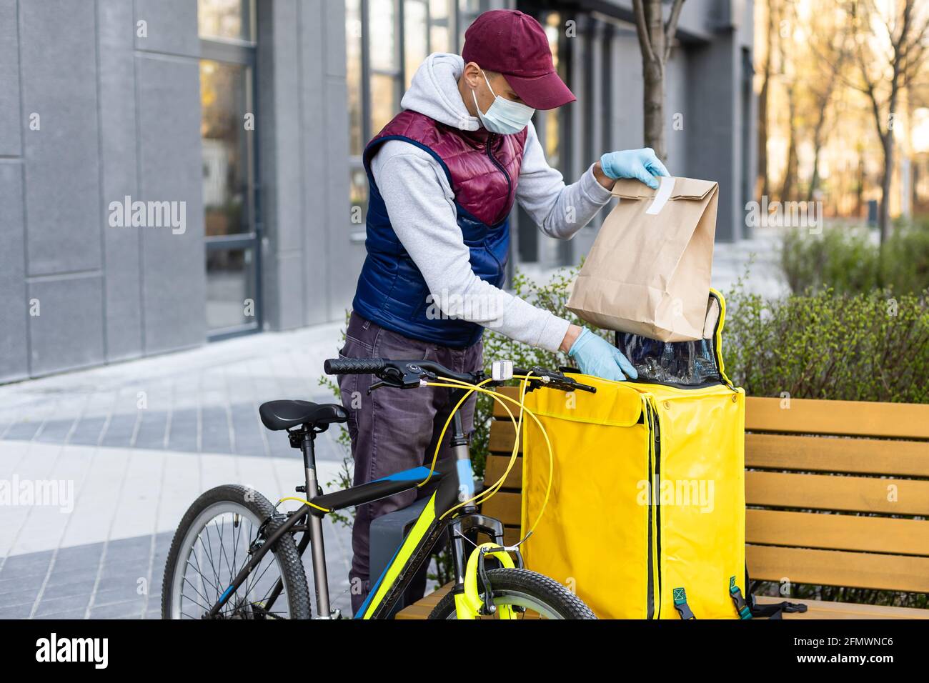 Junger Kurier in medizinischer Maske liefert Lebensmittel mit gelbem Thermalrucksack, fährt mit dem Fahrrad in der Stadt. Konzept für den Lieferservice von Lebensmitteln Stockfoto