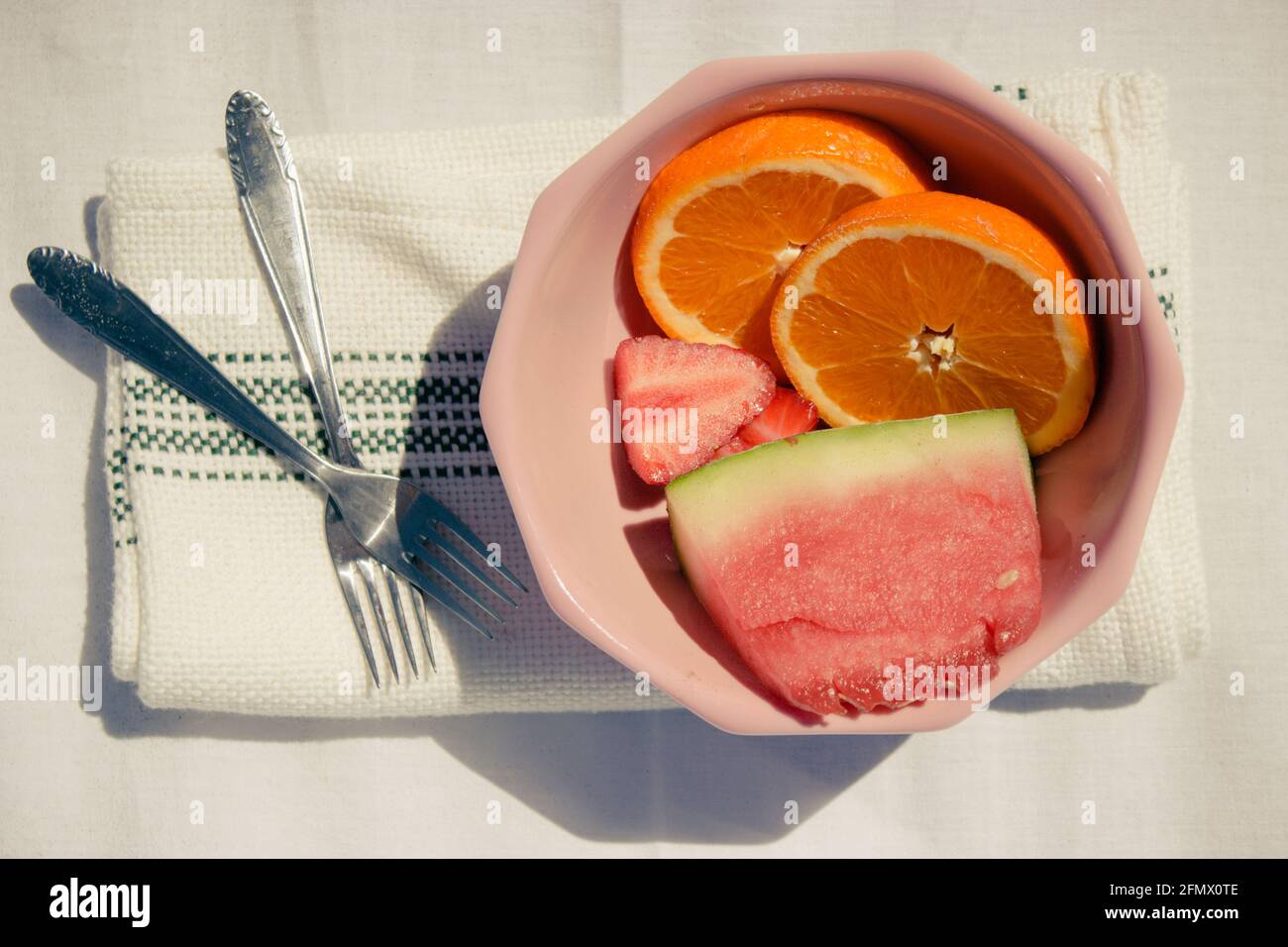 Detail von verschiedenen Früchten (Orangen, Erdbeeren und Wassermelone) in einer rosa Schüssel zusammen mit Gabeln und Handtuch auf einem weißen Stoffhintergrund. Sommer 2021 Stockfoto