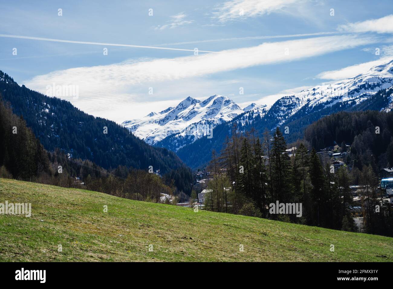 Blick von einem Grasfeld auf schneebedeckte Berge in St.Anton, Österreich. Coniefers und andere Bäume lassen die Szene grün und schön aussehen. Dorf Stockfoto