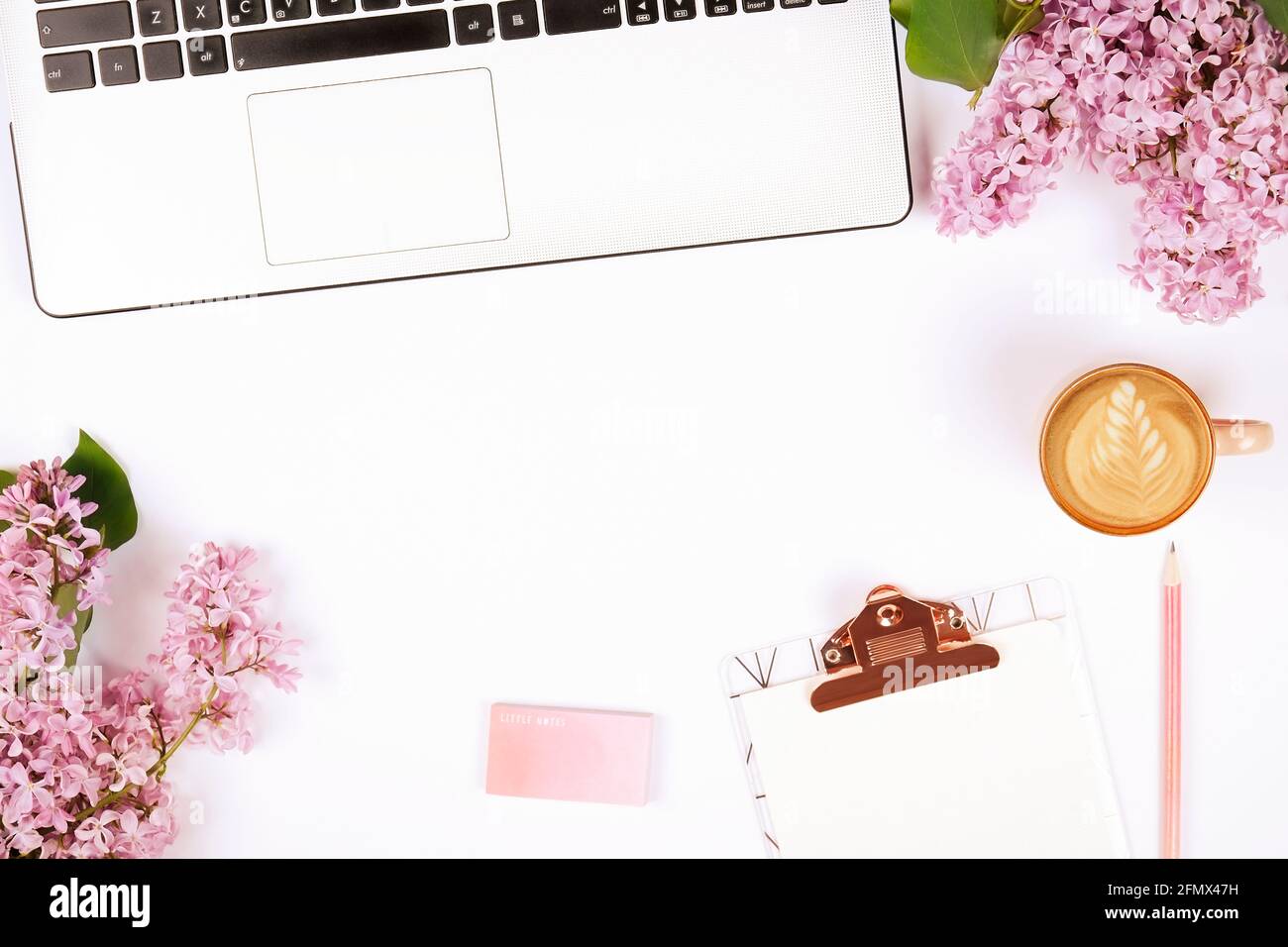 Feminine Nahaufnahme der Laptop-Computer-Tastatur, Kaffeetasse und lila Blumen. Flache Lay-Komposition mit Notizbuch, leeres Blatt-Klemmbrett, Radierer, pu Stockfoto