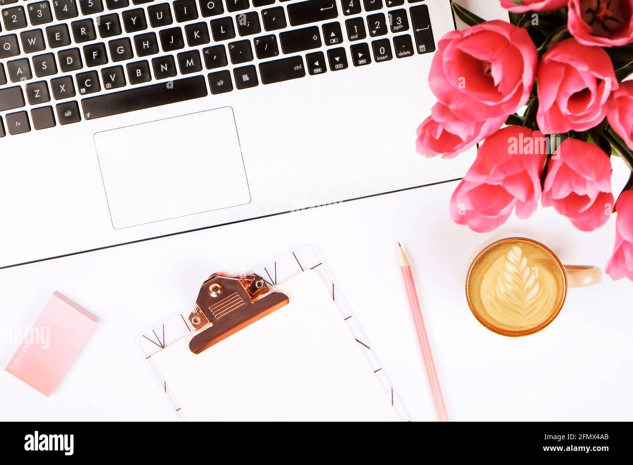 Feminine Nahaufnahme der Laptop-Computer-Tastatur, Kaffeetasse und rosa Tulpenblüten. Flache Lay-Komposition mit Notizbuch, leeres Blatt-Clipboar Stockfoto
