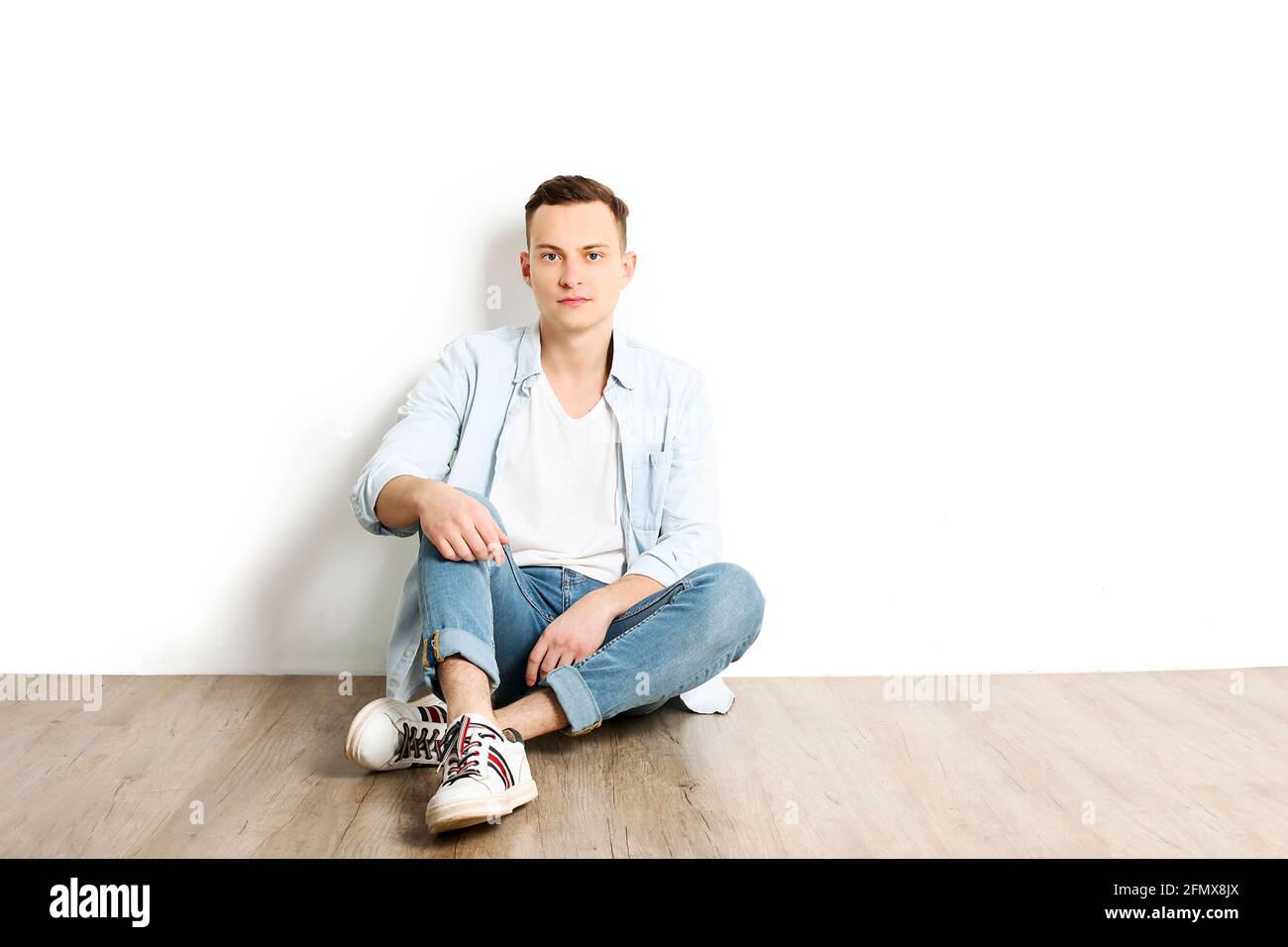 Porträt eines glücklichen jungen Millennials mit strahlenden Lächeln in einem weißen Hemd ohne Knöpfe sitzt auf einem Holzboden, isoliert. Selbstbewusster lässiger Hippie-Look Stockfoto