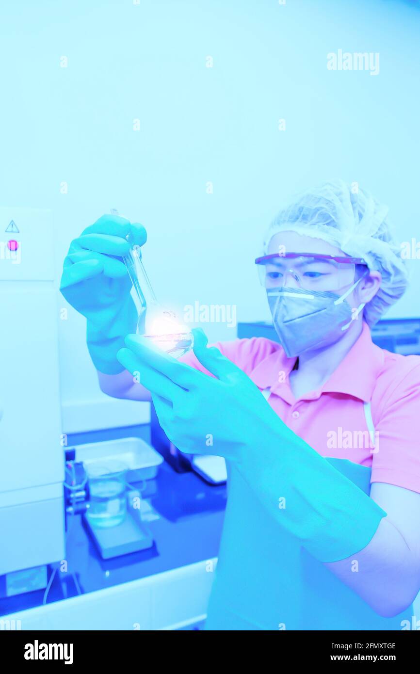 Eine asiatische Forscherin, die eine Operationsmaske mit einem volumetrischen Kolben oder Reagenzglas trägt, um in einem Labor zu forschen. Stockfoto