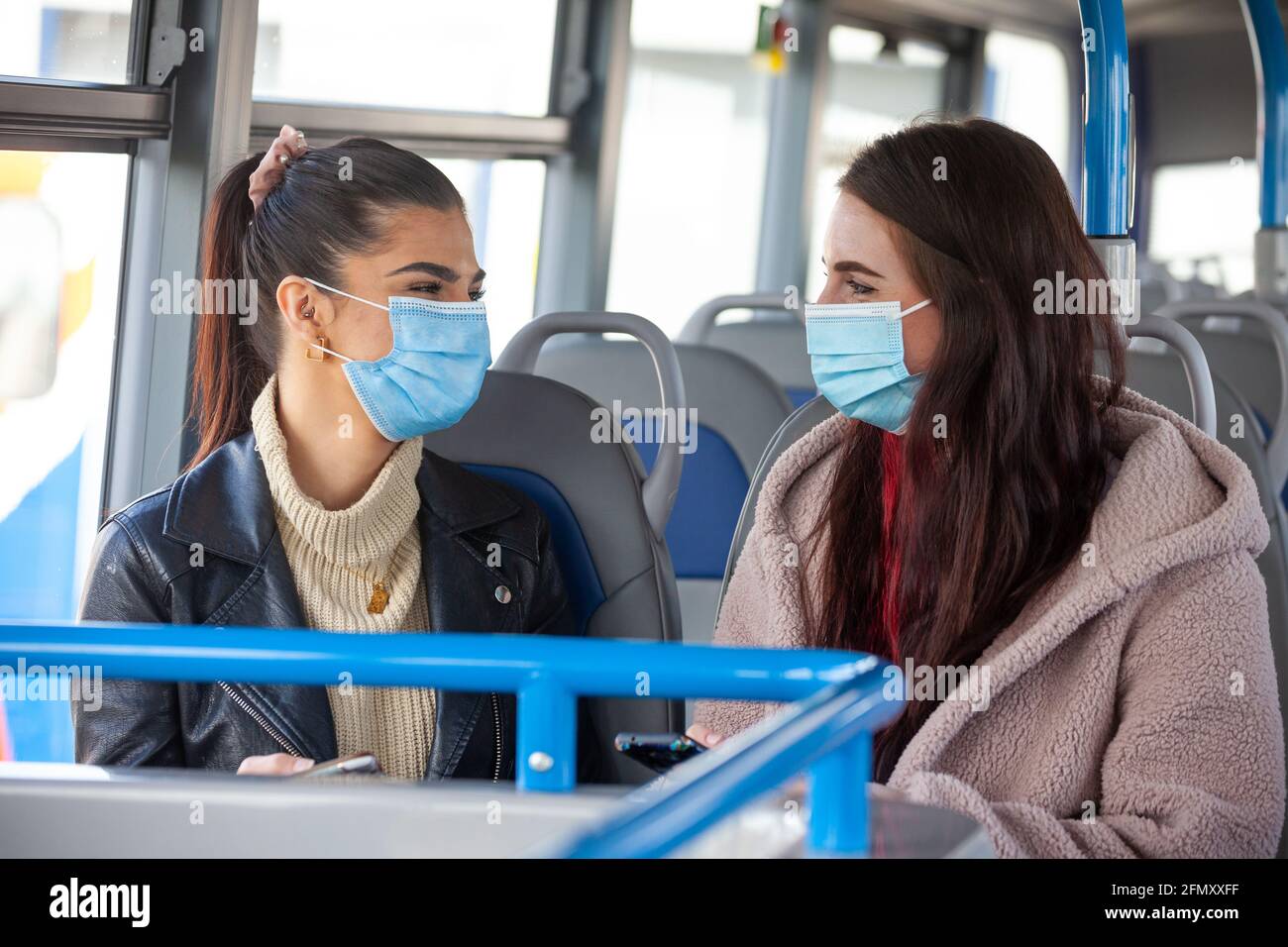 Zwei Frauen in ihren zwanzig Jahren tragen Gesichtsmasken und unterhalten sich mit ihnen Jeder in einem Bus Stockfoto