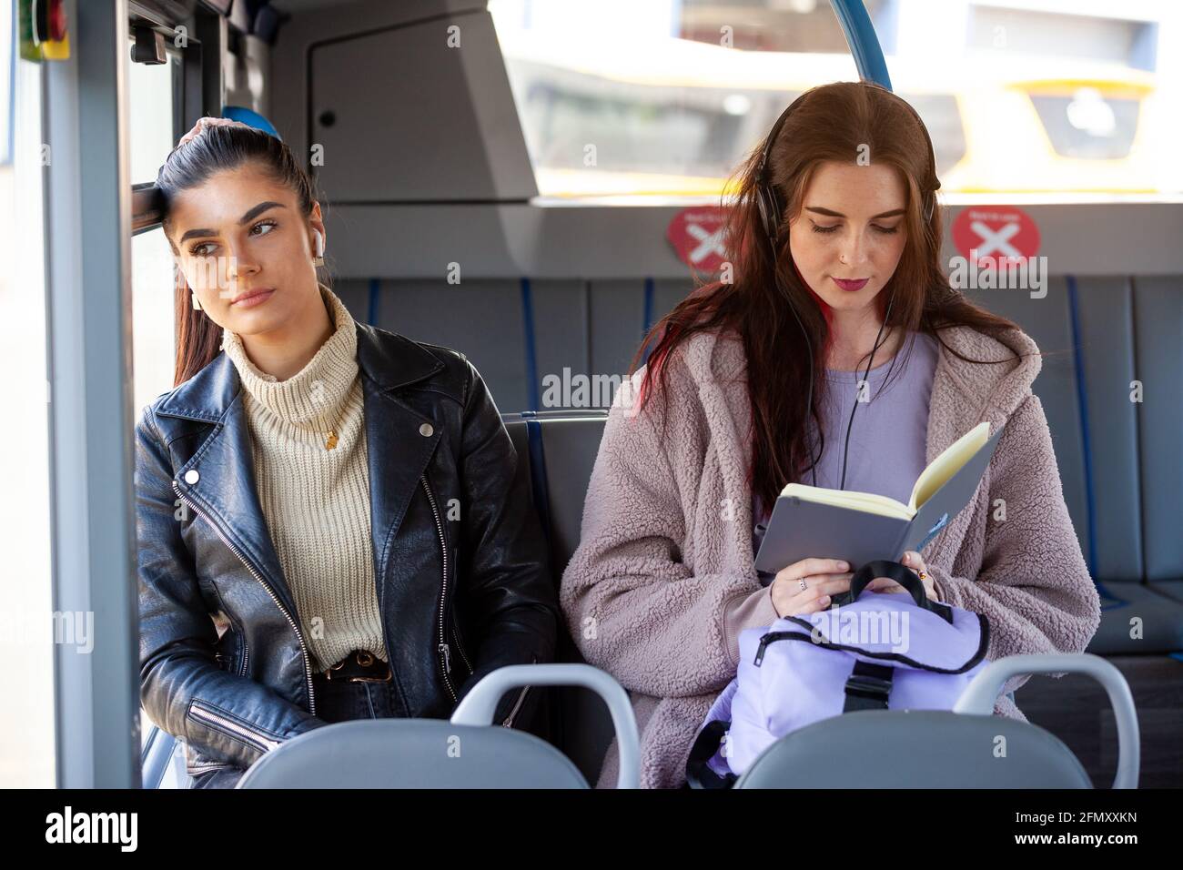 Zwei Frauen mit Schutzmasken sitzen auf einem Bus Einer schaut nebeneinander aus dem Fenster und der andere Lesen Stockfoto
