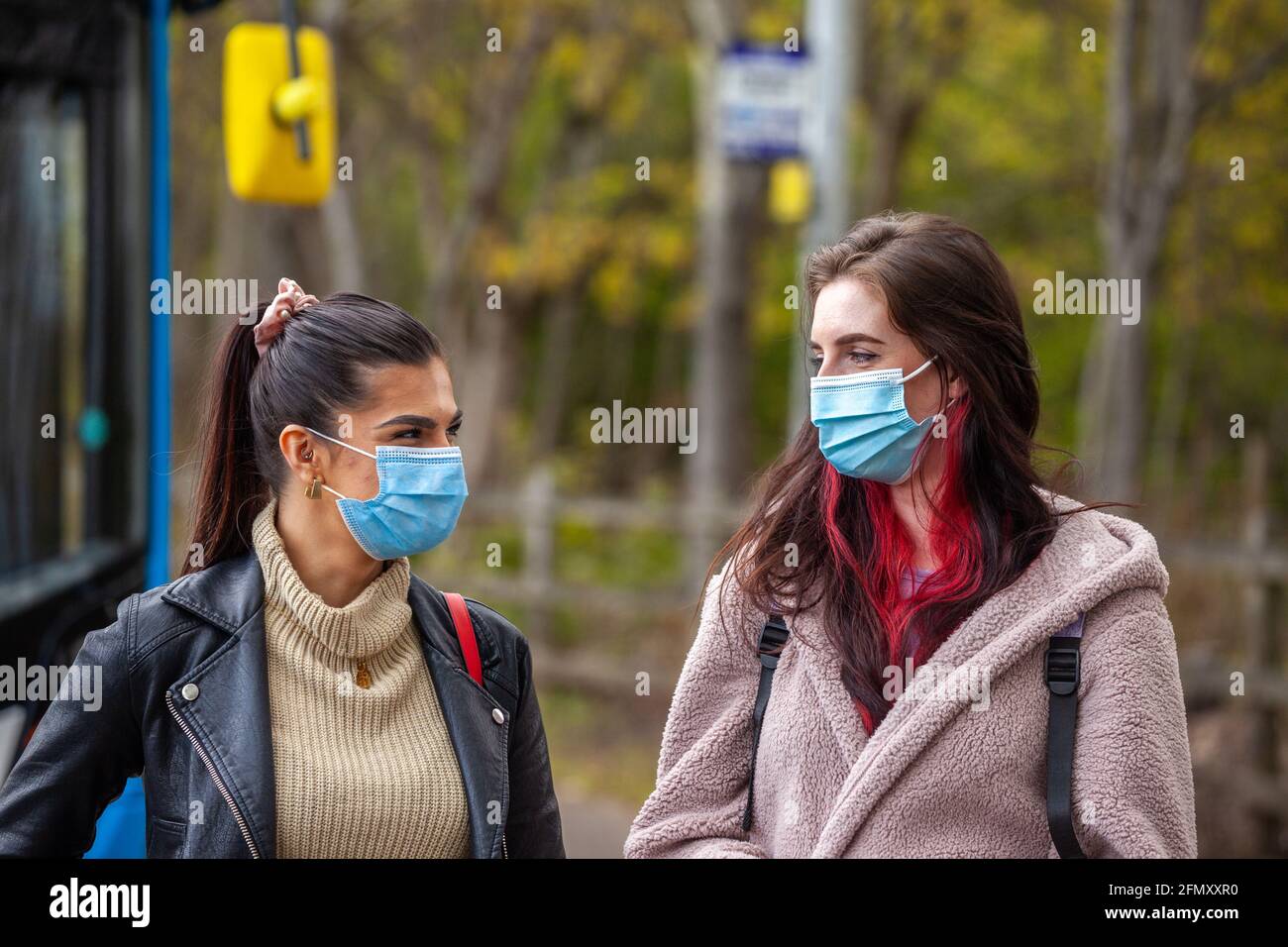 Zwei junge Frauen mit Gesichtsbezügen warten vor einem Bus an einer Bushaltestelle. Stockfoto