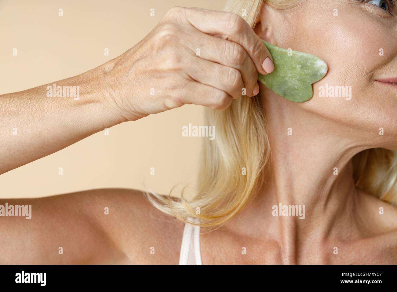 Nahaufnahme einer reifen blonden Frau mit perfektem Leuchten Haut mit Jade Gua Sha Massagewerkzeug für die Hautpflege Und Schönheitsbehandlungen isoliert über Beige Stockfoto