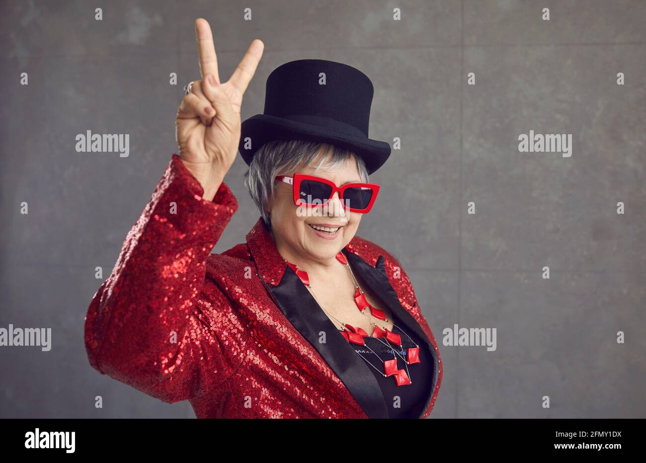Glückliche ältere Frau in Pailletten-Outfit, Hut und coole Brille tun V Zeichen Geste Stockfoto