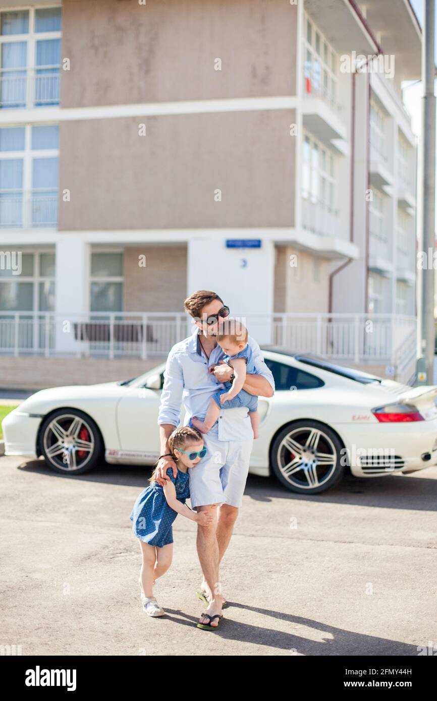 Der junge Vater läuft mit seinen beiden Töchtern auf dem Hintergrund eines Sportwagens Stockfoto