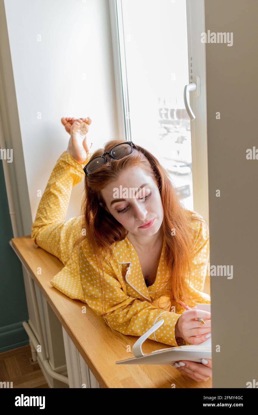 Eine junge Frau im bequemen gelben Pyjama arbeitet und liegt auf der Fensterbank im Schlafzimmer. Geschäftsfrau unterstützt soziale Distanzierung, arbeitet aus der Ferne. Neues normales Konzept Stockfoto