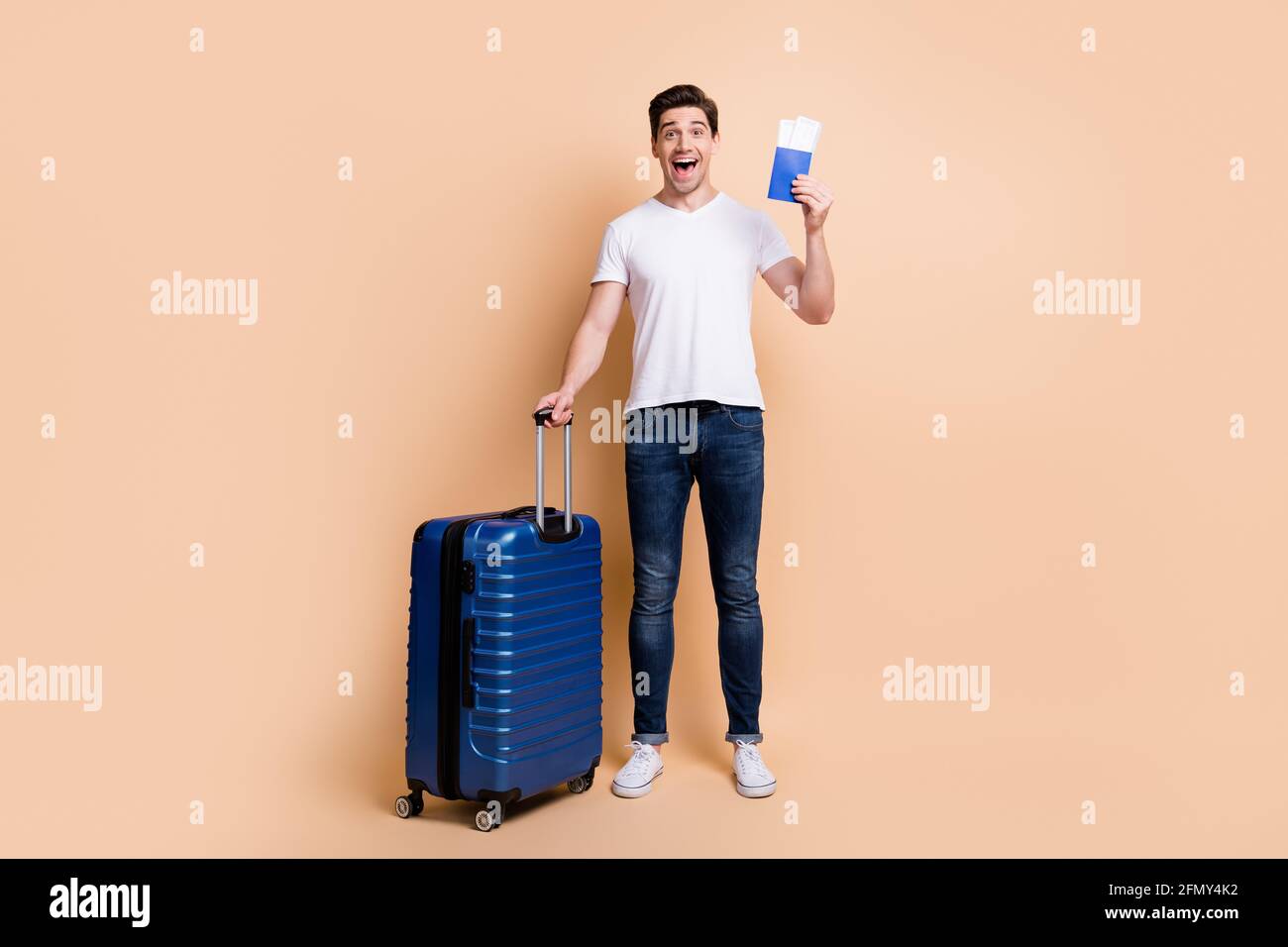 Foto in voller Größe von beeindruckt Brunet Mann Stand halten Tickets Tasche tragen T-Shirt Jeans Sneakers isoliert auf beigem Hintergrund. Stockfoto