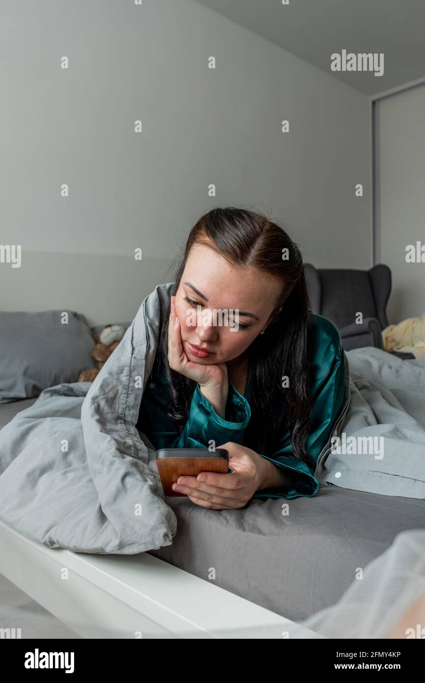 Der Seidenschlafanzug der jungen Frau, der im Bett liegt, hält ihr Handy an den Händen. Arbeiten Sie bequem von zu Hause aus. Wellness-Konzept. Stockfoto