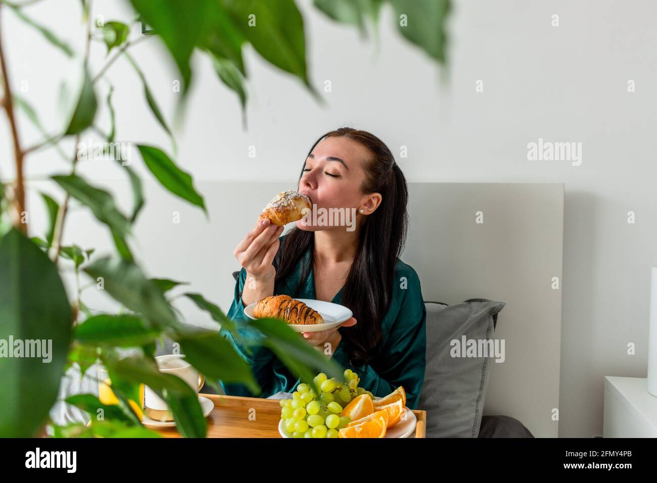 Die junge Frau im Seiden-Pyjama hat ein gesundes Frühstück, während sie im Schlafzimmer auf dem Bett liegt. Konzept für das Wohlbefinden zu Hause. Emotionale Gesundheit einer jungen Frau Stockfoto