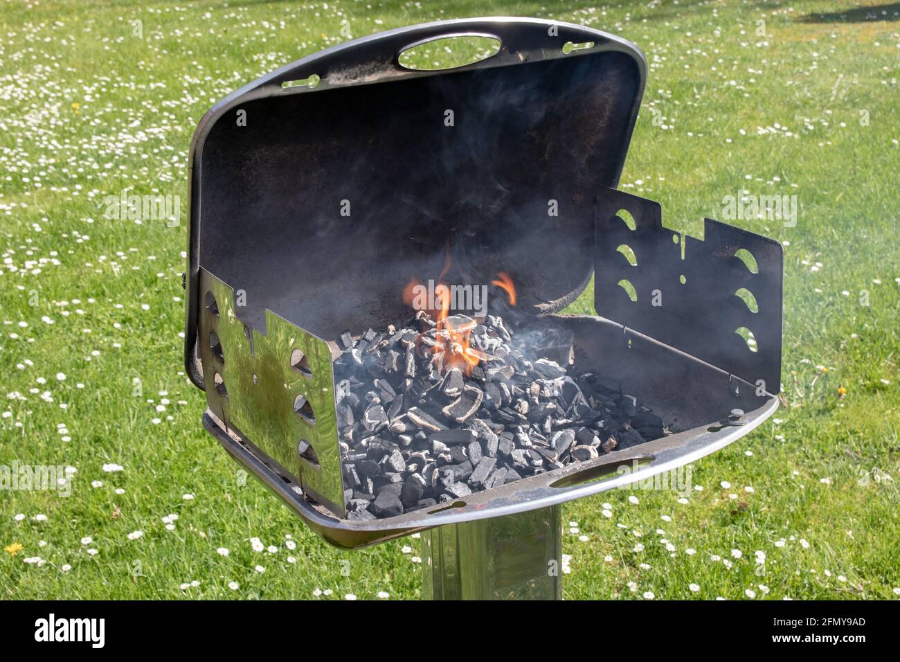 Ein heißer brennender, leerer Holzkohlegrill mit offenem Feuer. An einem sonnigen Frühlingstag wird auf einer grünen Wiese mit Gänseblümchen ein köstliches Barbecue im Freien vorbereitet. BBQ Stockfoto