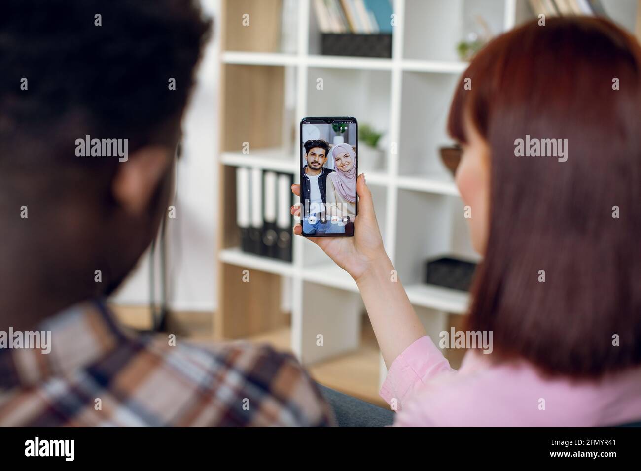 Betrachten Sie von der Schulter eines multirassischen Paares, das mit einem modernen Smartphone in den Händen auf der Couch sitzt und mit muslimischen Freunden per Videoanruf spricht. Konzept von Mensch und Technik. Stockfoto