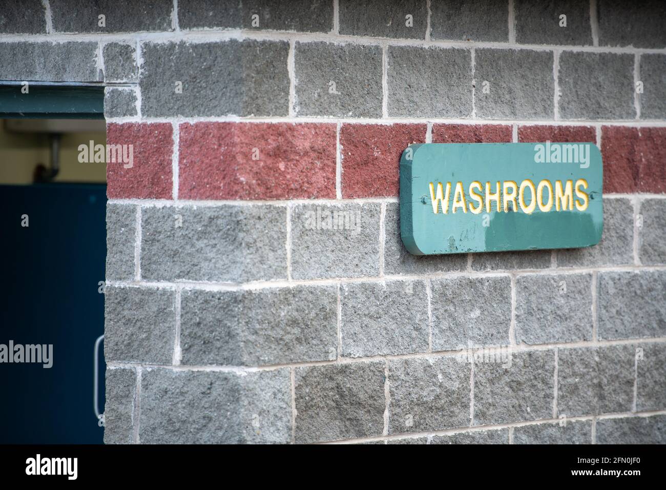 Ein Waschraum-Schild mit gelben Buchstaben auf grünem Hintergrund in einem Park. Das Schild ist grün. Die Wand ist aus grauem Ziegelstein mit einem roten Ziegelstreifen. Stockfoto