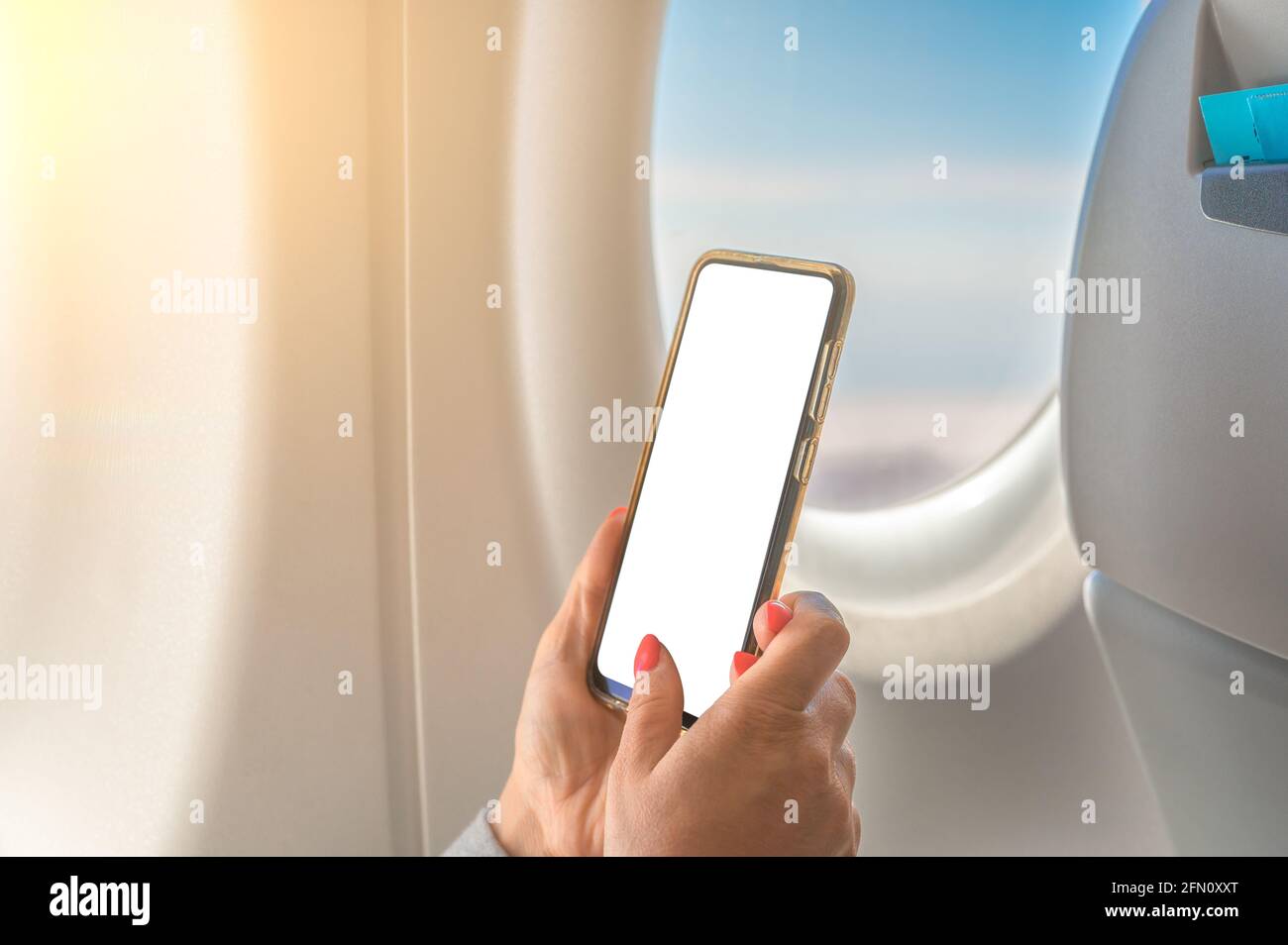 Mockup-Bild einer Frau, die am Smartphone mit leerem weißen Bildschirm hält, Flugzeugfenster mit Himmelshintergrund. Hand mit Handy im Flugzeug. Weiblich Stockfoto