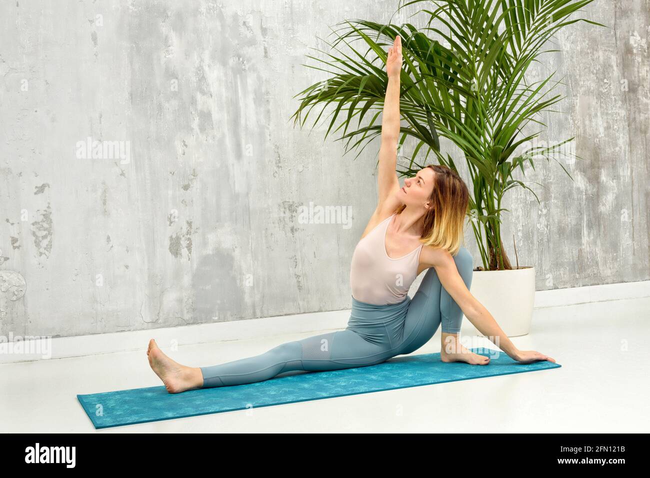 Frau, die eine Upavista Konasana Variation Yoga Pose macht, die sie dehnt Arm über ihrem Kopf mit einer grauen Wand und vergossen Pflanze Hintergrund in einer Gesundheit und fitn Stockfoto