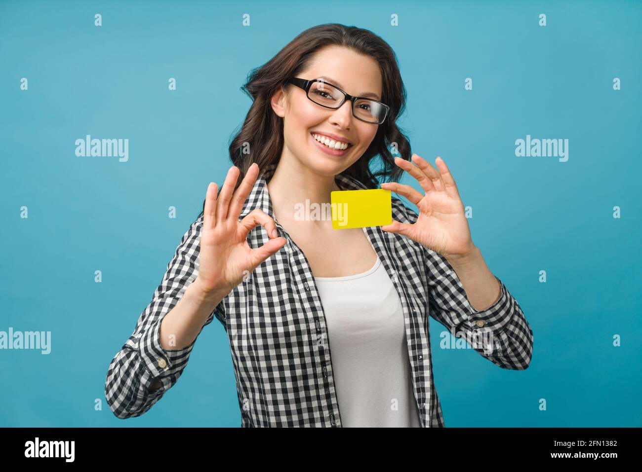 Niedliche lächelnde Frau in Hemd und Brille, zeigt Kreditkarte in der Hand für Finanz- und bargeldlose Gesellschaft Konzept, posiert auf blauem Hintergrund Stockfoto