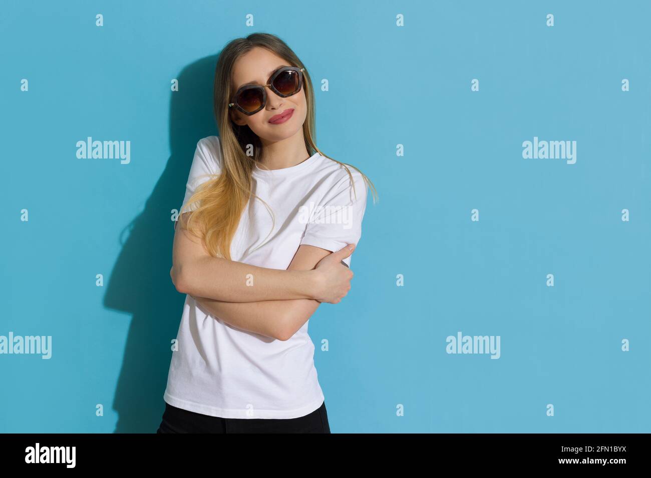 Glückliche junge Frau in weißem Hemd und Sonnenbrille hält gekreuzte Arme, schaut zur Seite und lächelt. Studio-Aufnahme auf blauem Hintergrund mit Hüfthöhe. Stockfoto