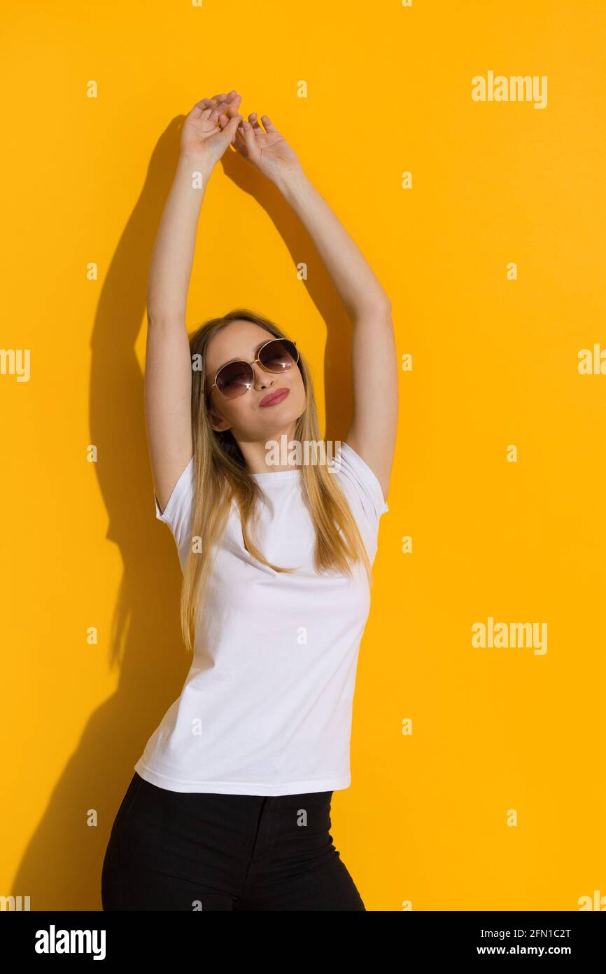 Glückliche junge blonde Frau in weißem Hemd und Sonnenbrille genießt Sonnenlicht an der gelben Wand. Stuido-Aufnahme in der Taille. Stockfoto