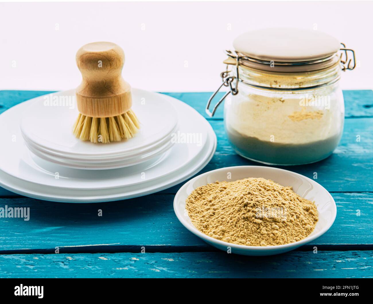 Verwendung von Senfpulver zum Waschen des Geschirrkonzepts. Weiße saubere Teller mit Schale aus gelbem Senfpulver und Holzwaschbürste und Klassenglas Stockfoto