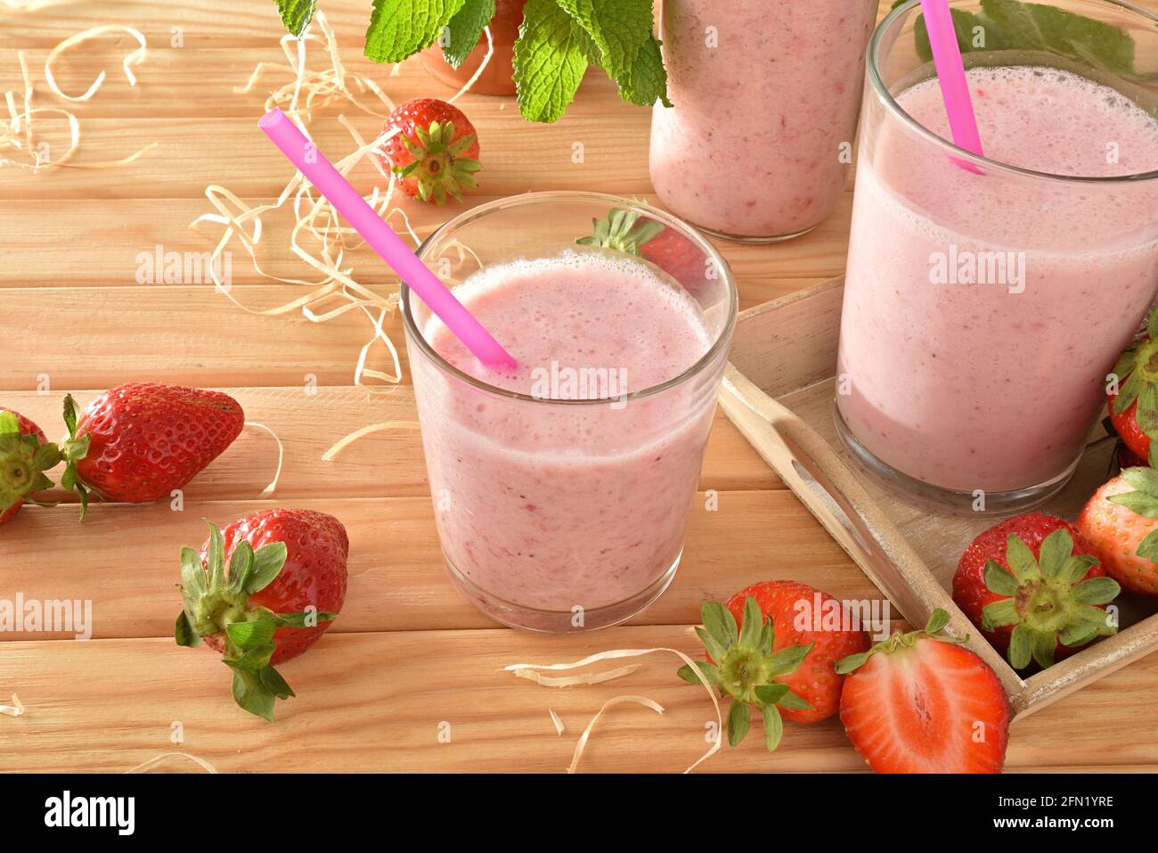 Erdbeer-Smoothie mit Milch in Glasbehältern auf Holztisch mit Tablett voller Erdbeeren in der Natur. Erhöhte Ansicht. Stockfoto