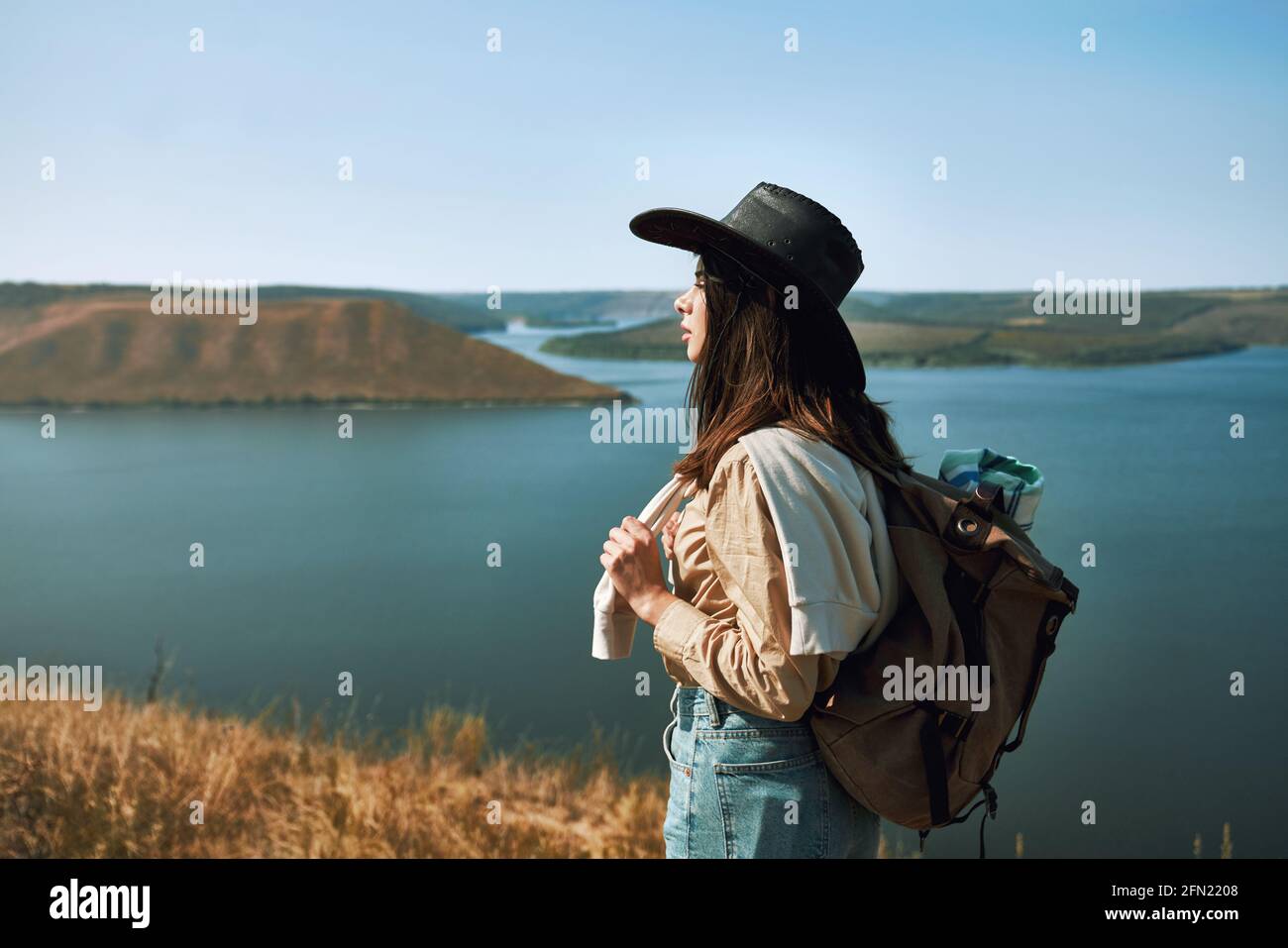 Positive junge Frau im Cowboyhut, die in der Gegend von Bakota durch die grüne Natur spazierengeht. Glückliche Hündin mit Rucksack verbringen Freizeit aktiv. Stockfoto