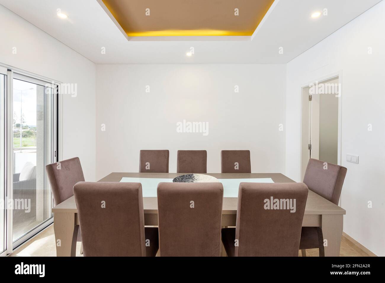 Gut beleuchteter Speisesaal mit einem schönen modernen Tisch und acht Stühlen um ihn herum, es gibt eine Tür zu einem anderen Raum und ein Fenster nach außen. Stockfoto