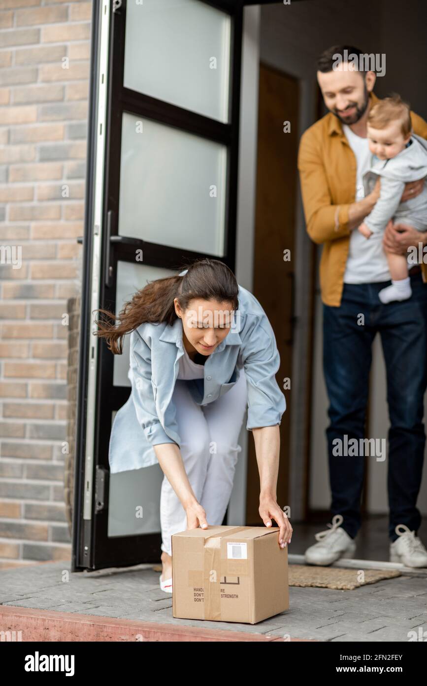 Junge Eltern mit neugeborenen Kindern bestellen die Paketzustellung online und stehen vor dem Haus, um sie zu erhalten. Hausfrau bringt ein Paket, während Ehemann ein Baby hält. Stockfoto