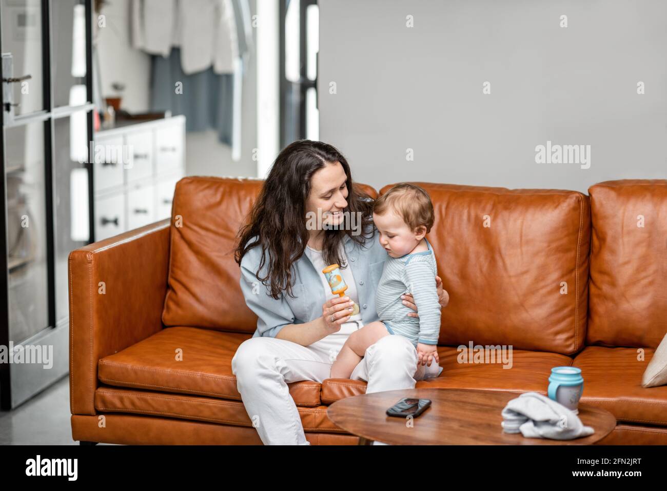 Junge Mutter sitzt mit neugeborenen Sohn auf dem Sofa und kümmern oder spielen mit einem Kind. Aufzucht von Baby, Babysitter und Mutterschaft. Stockfoto