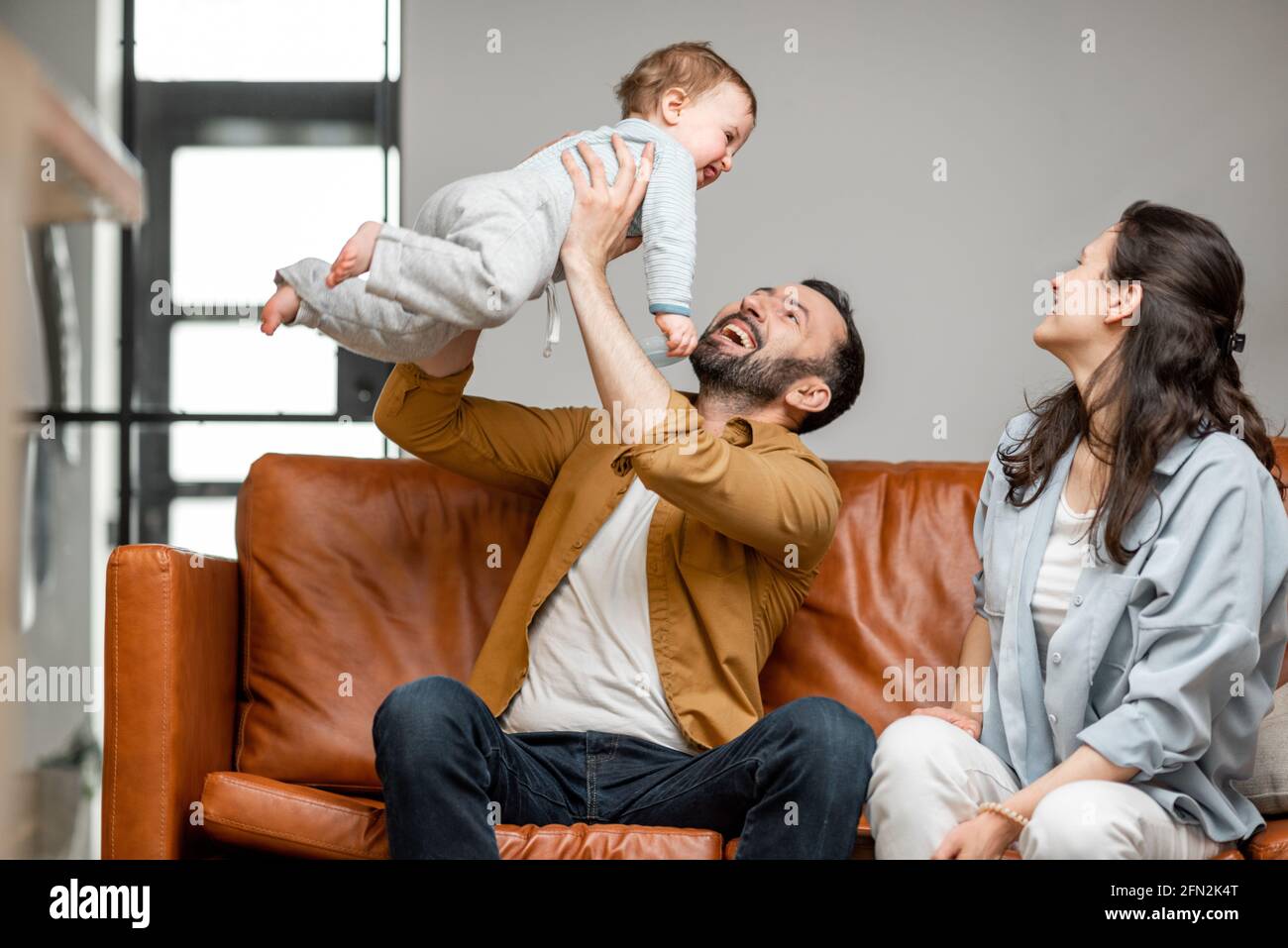 Glücklicher Vater, Mutter und kleiner Sohn spielen auf dem Sofa im Wohnzimmer. Kinderbetreuung und Elternschaft. Papa wirft das Baby hoch. Stockfoto