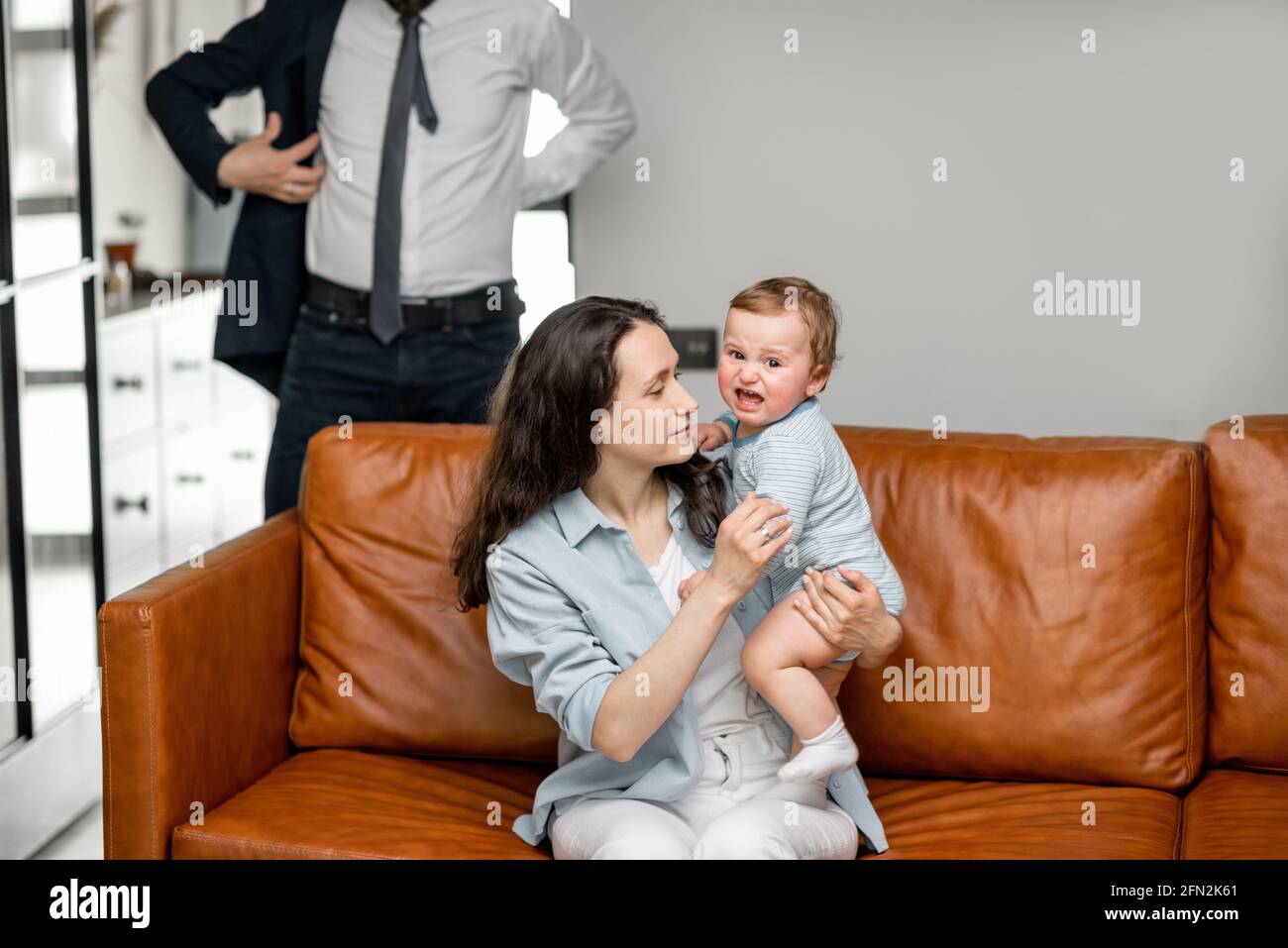 Junge Mutter sitzt mit neugeborenen Sohn auf dem Sofa und kümmern oder spielen mit einem Kind, während Vater und Mann zur Arbeit gehen und sagen Auf Wiedersehen. Stockfoto