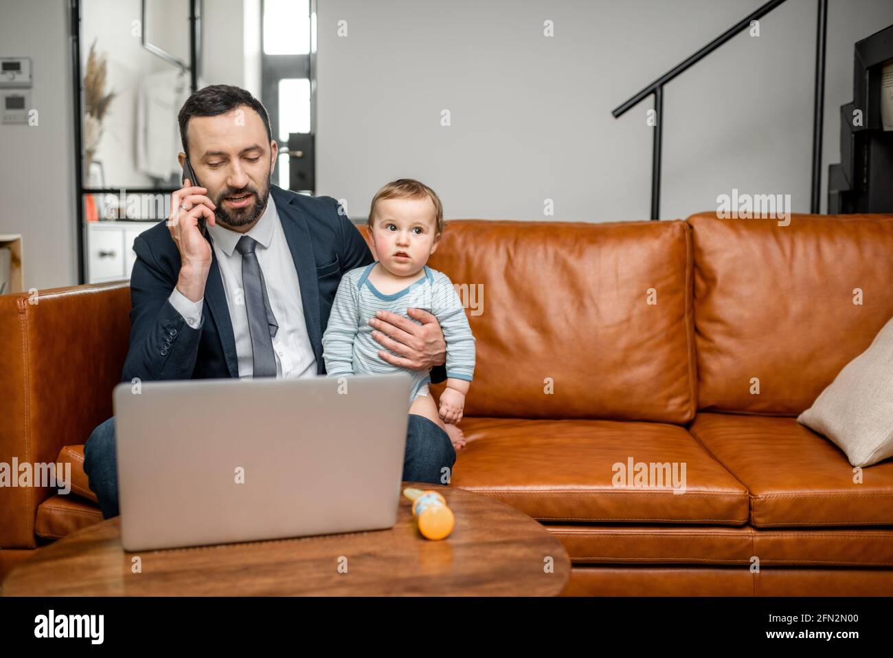 Junger Vater, der an einem Laptop arbeitet und zu Hause telefoniert, während er sich um seinen kleinen Sohn kümmert. Multitasking und Babysitter. Stockfoto