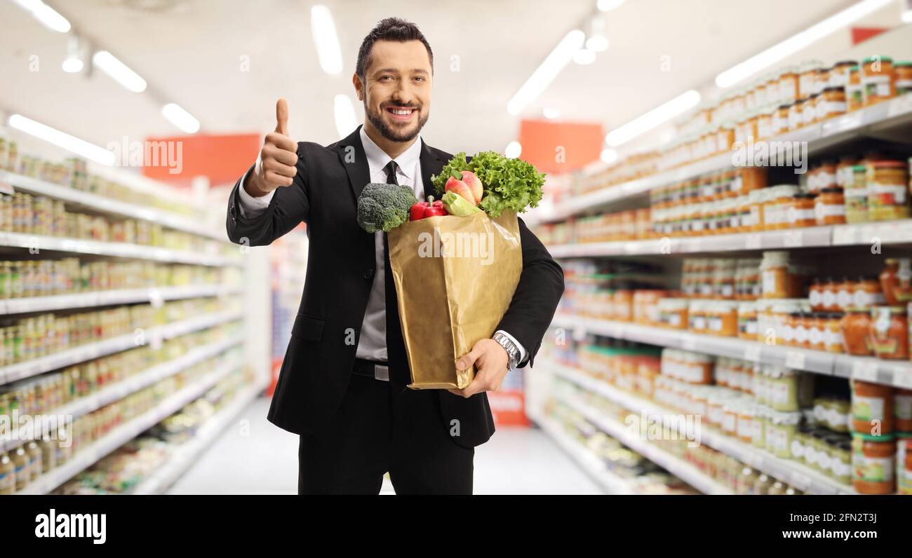 Junger Geschäftsmann mit einer Lebensmitteltasche, die in einem Supermarkt posiert Und ein Daumen hoch Zeichen gestikulieren Stockfoto