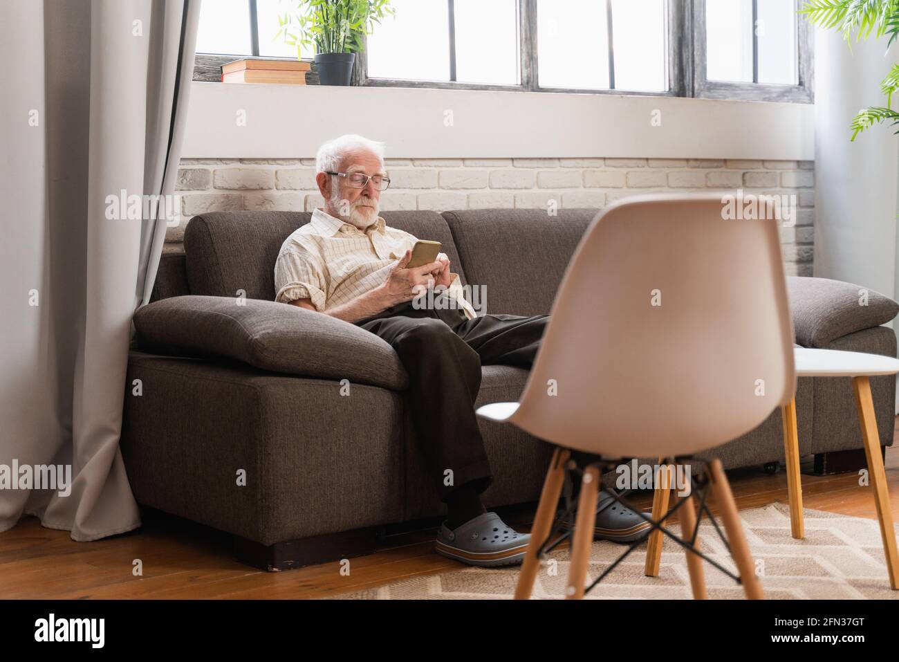 Älterer Mann sitzt auf dem Sofa und schreibt auf einem modernen Smartphone - Alter Mann allein zu Hause, Video ruft seine Familie per Mobiltelefon an Stockfoto