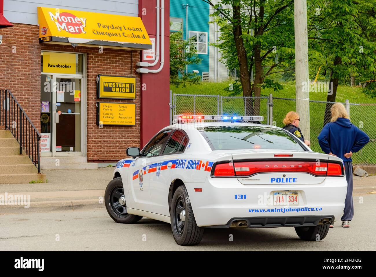 Saint John, NB, Kanada - 1. Juli 2015: Ein Polizeiauto blockiert die Straße vor einem Geldmarkt, während eine Polizistin mit einer Zuschauerin spricht. Stockfoto