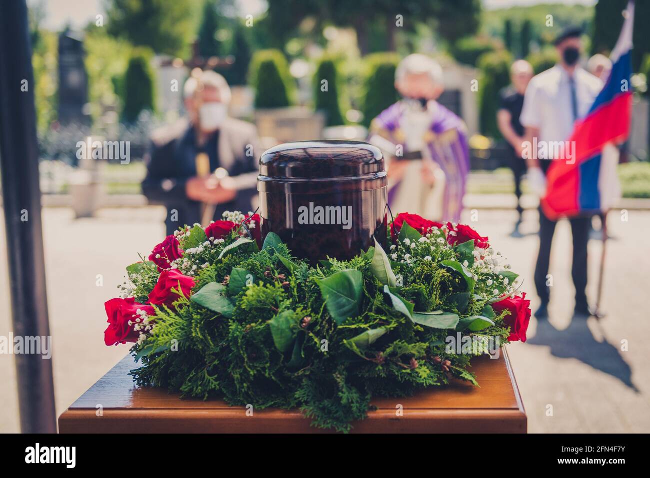 Begräbnisurne mit Asche von Toten und Blumen bei der Beerdigung. Begräbnisurne mit Blumen und chatolischen christlichen Priester im Hintergrund bei Memorial serv Stockfoto