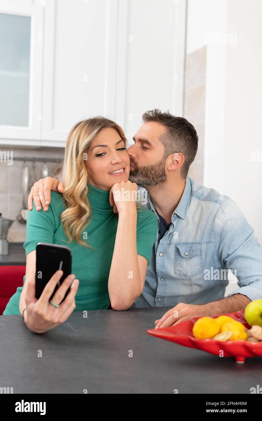 Die blonde Frau macht ein Selfie, während sie von ihrem Freund geküsst wird. Mädchen mit grünem Hemd fotografiert sich für soziale Medien. Glückliches Paar. Stockfoto