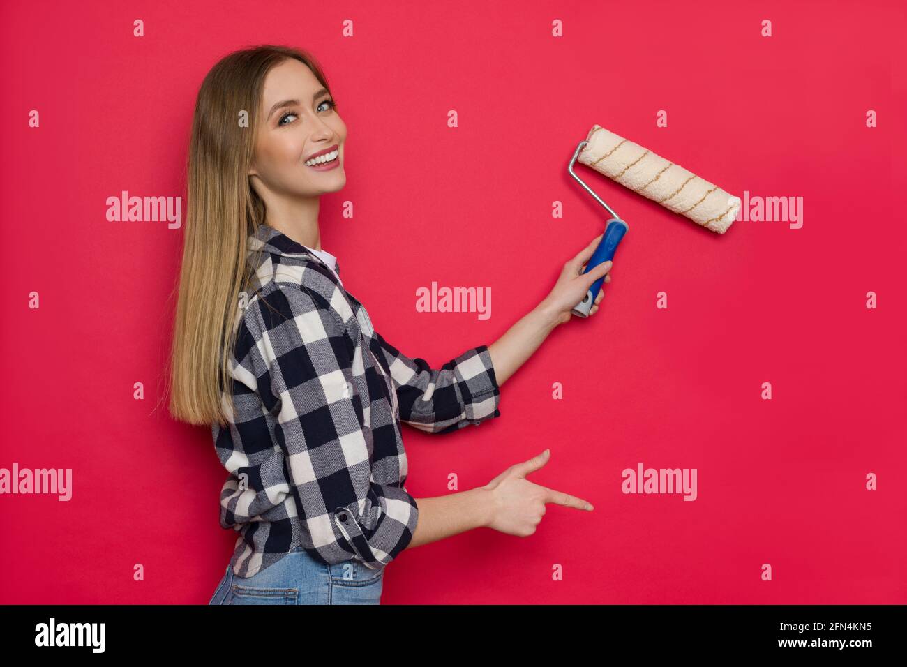 Die junge blonde Frau in Holzfällerhemd und Jeans malt mit Malrolle rote Wände, blickt über die Schulter auf die Kamera und zeigt. Taille nach oben Stockfoto