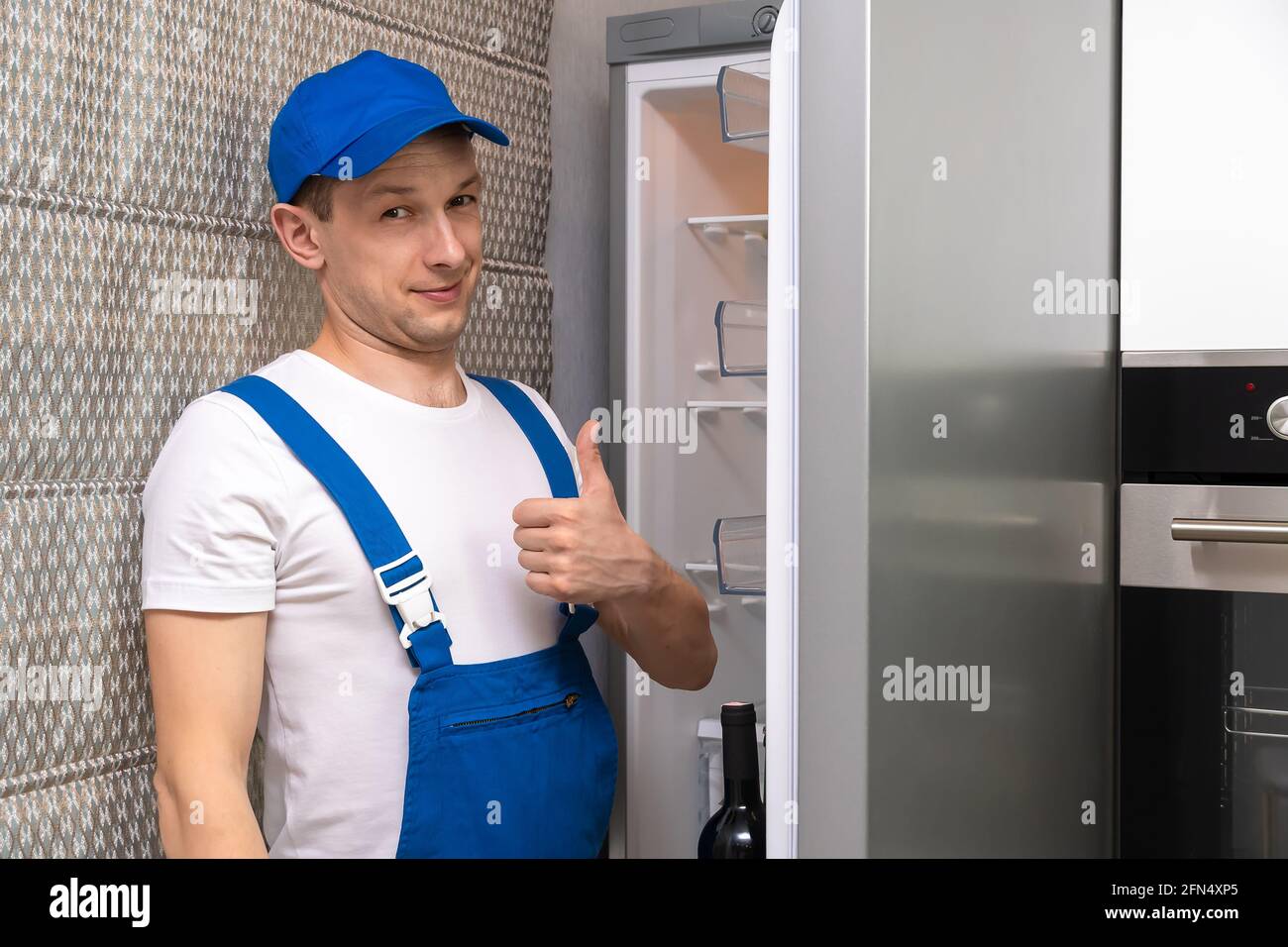 Uniformierte Werkstatt lächelt, schaut auf die Kamera und gibt vor dem Hintergrund eines offenen Kühlschranks in der Küche ein Daumen-nach-oben-Zeichen Stockfoto