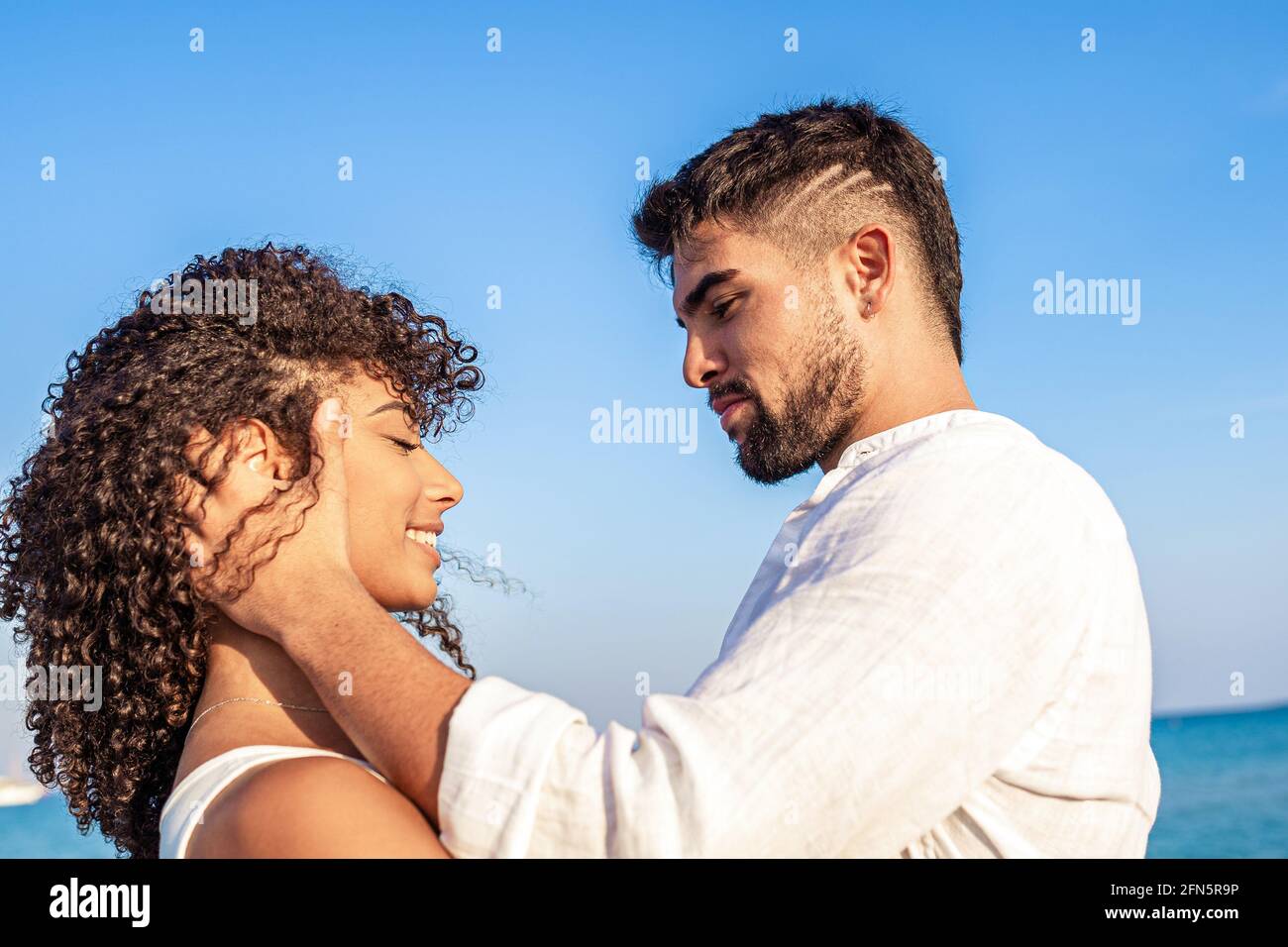 Moment der Zärtlichkeit zwischen jungen, multirassischen Paaren glücklicher Liebhaber, die einander in die Augen schauen, während er lächelnd ihren Kopf in den Händen hält. Bestanden Stockfoto