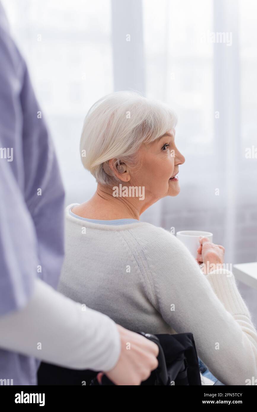 Behinderte ältere Frau mit Hörgerät, die eine Tasse Tee hält In der Nähe von Sozialarbeitern auf verschwommenem Vordergrund Stockfoto