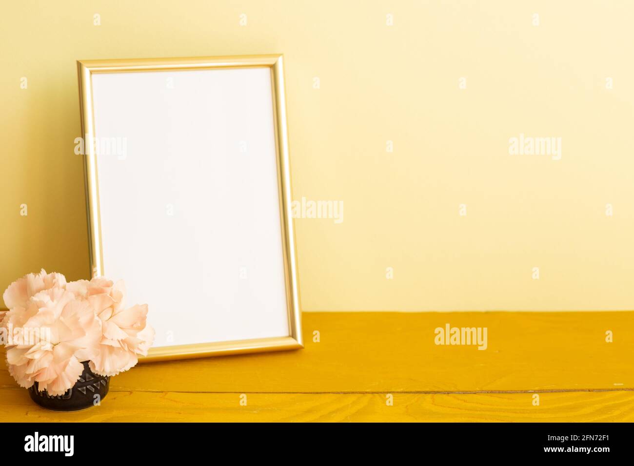 Leerer Bilderrahmen mit Nelkenblumen auf Holztisch. Gelber Hintergrund Stockfoto