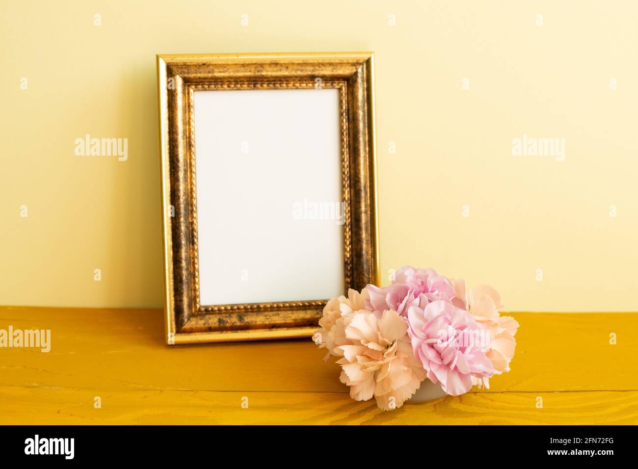 Leerer Bilderrahmen mit Nelkenblumen auf Holztisch. Gelber Hintergrund Stockfoto