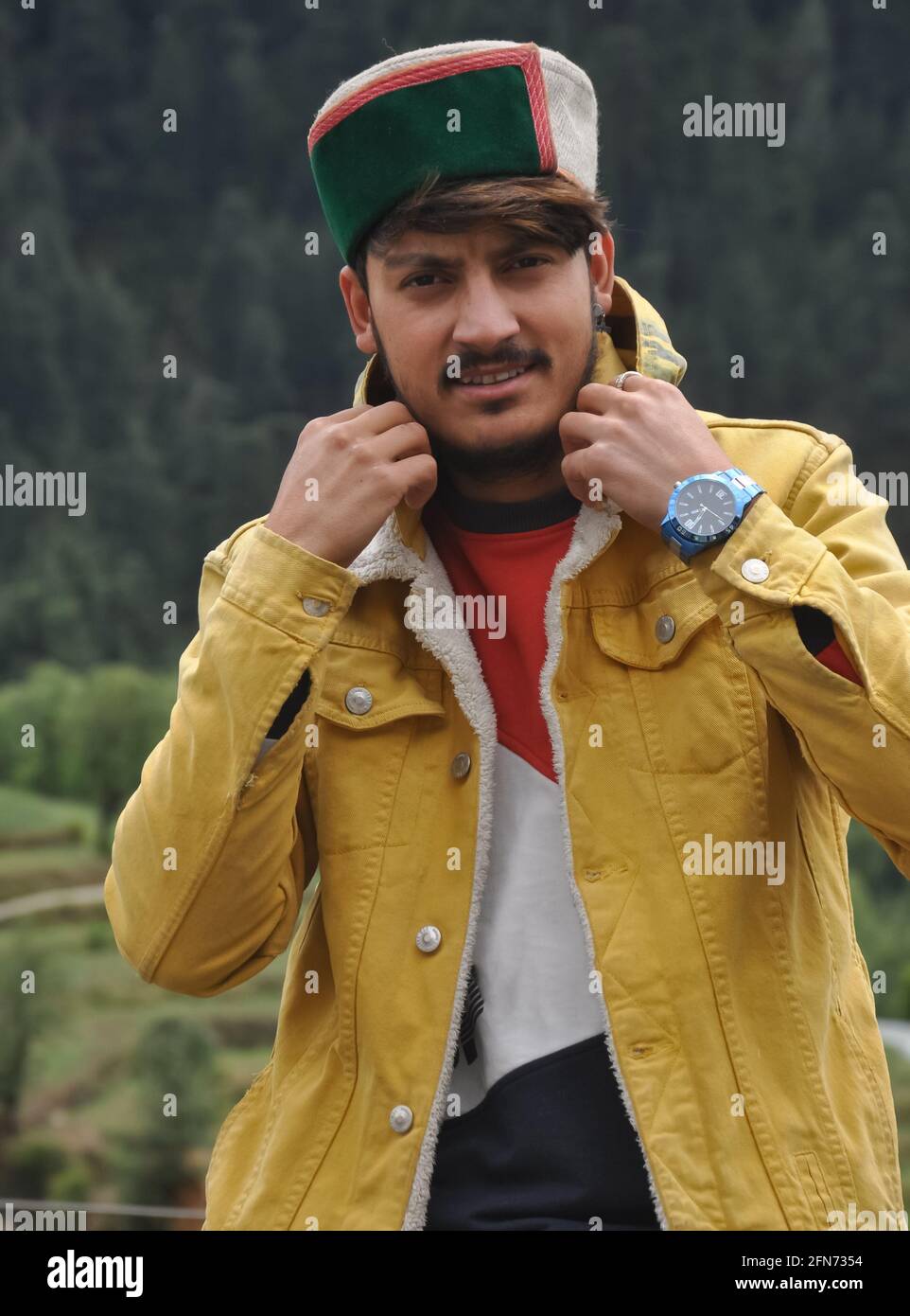 Porträt eines hübschen jungen Mannes, der seinen Jackenkragen anpasst, eines gut aussehenden jungen Mannes mit gelber Jacke und himachali topi (traditionelle Himachal Mütze) Stockfoto