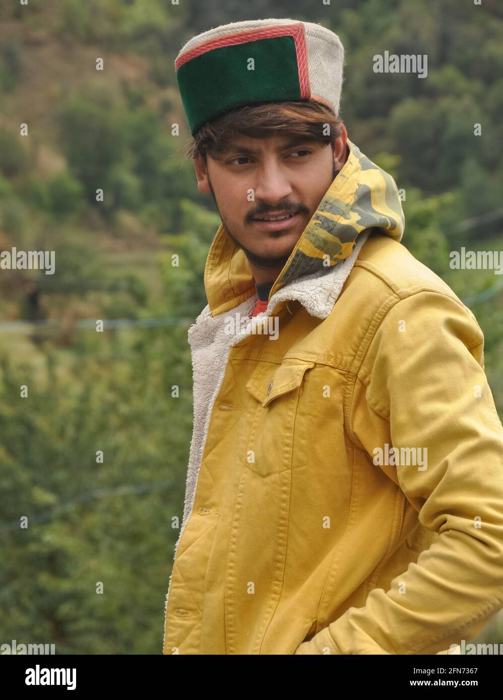 Seitenansicht eines gutaussehenden Mannes mit der Hand auf der Tasche und seitlichem Blick, EINES gut aussehenden jungen Mannes mit gelber Jacke und himachali topi (Himachali traditionelle Mütze) Stockfoto